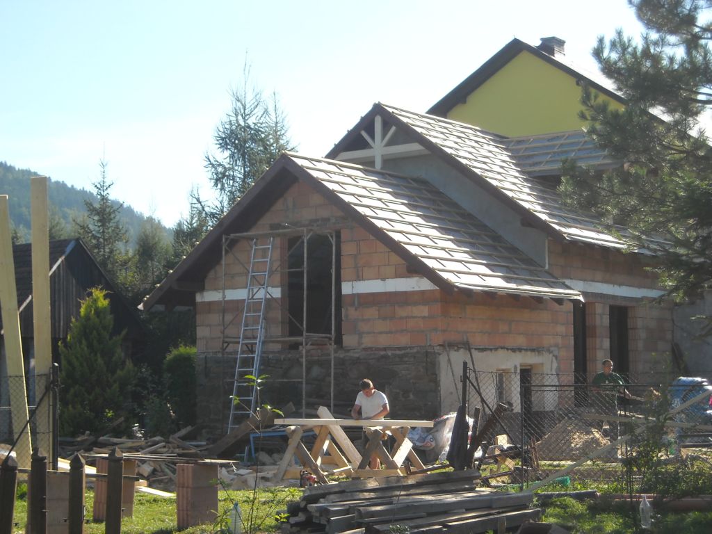 Wznoszenie murów domu z cegły ceramicznej, widoczne rusztowanie, przygotowane drewno i pracownicy na placu budowy.